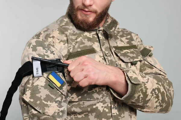 Ukrainian soldier in military uniform applying medical tourniquet on arm against light grey background, closeup