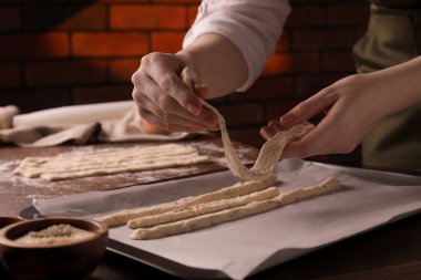 Woman putting homemade breadsticks on baking sheet at wooden table indoors, closeup. Cooking traditional grissini