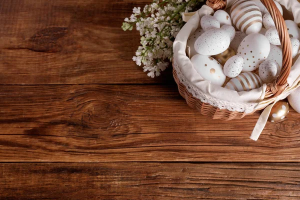 stock image Wicker basket with festively decorated Easter eggs and white lilac flowers on wooden table, flat lay. Space for text