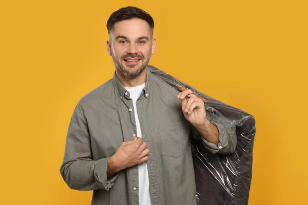Man holding garment cover with clothes on yellow background. Dry-cleaning service