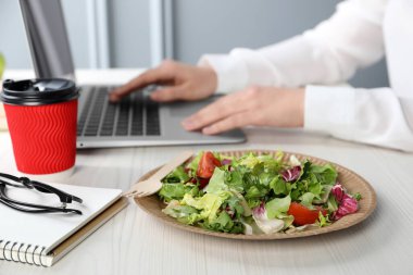 Beyaz ahşap masada dizüstü bilgisayarla çalışan bir ofis çalışanı, sebze salatası ve kağıt bardak kahveye odaklanıyor. İş yemeği.