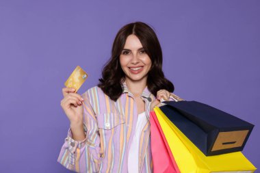 Güzel bir genç kadın, kağıt alışveriş torbaları ve mor arka planda kredi kartıyla.