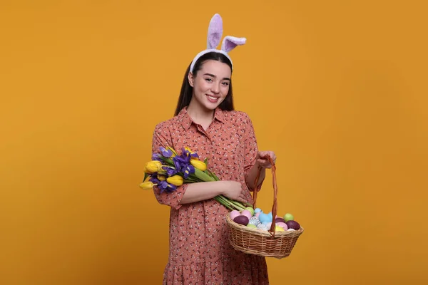 戴着兔子耳朵帽带的快乐女人拿着柳条篮 上面有彩绘的复活节彩蛋和橙色背景的花束 — 图库照片