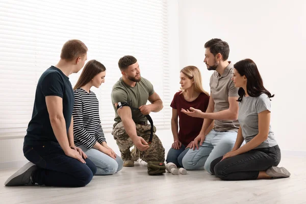 Soldier in military uniform teaching group of people how to apply medical tourniquet indoors