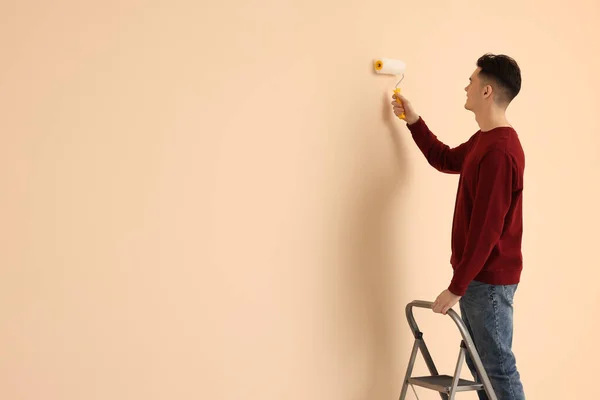 Young man painting wall with roller on stepladder indoors, space for text. Room renovation