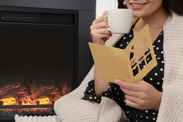 Young woman sitting near fireplace with greeting card and hot drink indoors, closeup. Space for text