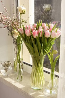 Many different spring flowers and branches with leaves on windowsill indoors