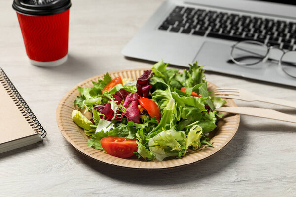 Fresh vegetable salad, laptop and paper cup of coffee on white wooden table at workplace. Business lunch