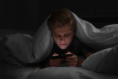 Genç çocuk gece yatakta battaniyenin altında akıllı telefon kullanıyor. İnternet bağımlılığı