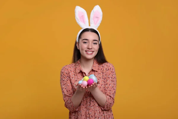 戴着兔子耳朵帽带的快乐女人 带着彩绘的复活节彩蛋 背景是橙色的 — 图库照片