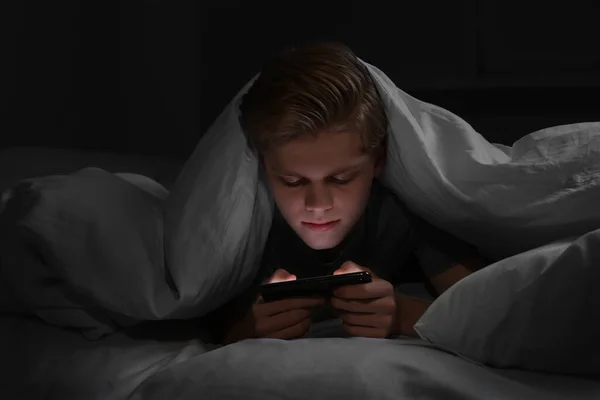 Teenage boy using smartphone under blanket on bed at night. Internet addiction