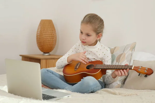 Little girl learning to play ukulele with online music course at home. Time for hobby