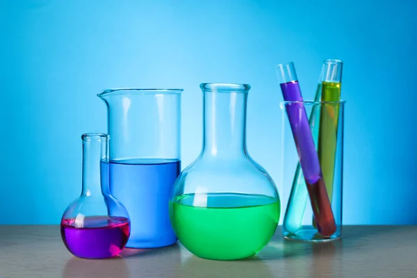 stock image Different laboratory glassware with colorful liquids on wooden table against light blue background