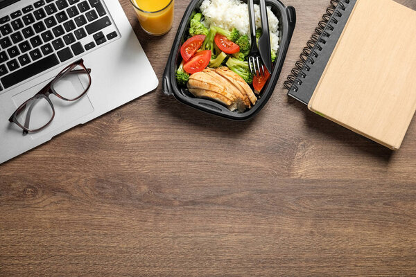 Container of tasty food, laptop, glasses and notebook on wooden table, flat lay with space for text. Business lunch