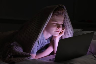 Genç çocuk geceleri yatakta battaniyenin altında dizüstü bilgisayar kullanıyor. İnternet bağımlılığı