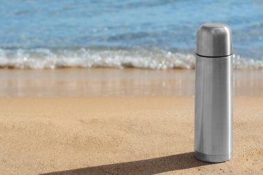 Metallic thermos with hot drink on sandy beach near sea, space for text