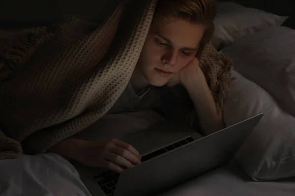 Teenage boy using laptop under blanket on bed at night. Internet addiction