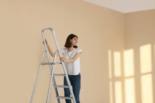 Young woman climbing up stepladder near wall indoors. Room renovation