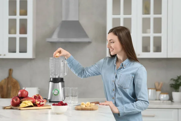 stock image Beautiful young woman adding banana into blender for tasty smoothie in kitchen