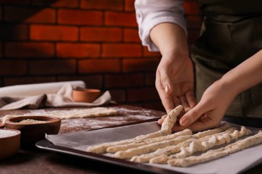 Woman putting homemade breadsticks on baking sheet at wooden table indoors, closeup. Cooking traditional grissini