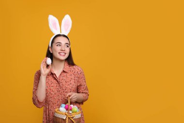 Happy woman in bunny ears headband holding wicker basket of painted Easter eggs on orange background. Space for text