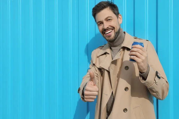 Handsome man holding tin can with beverage and showing thumbs up near light blue wall. Space for text