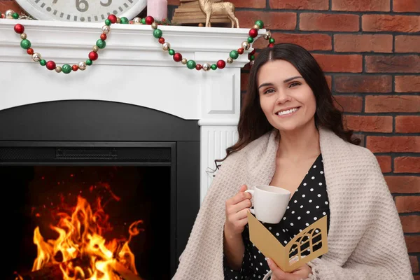 Young woman with greeting card and hot drink sitting near fireplace indoors
