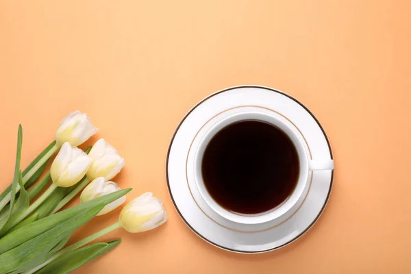 Cup of coffee and beautiful tulips on light orange background, flat lay