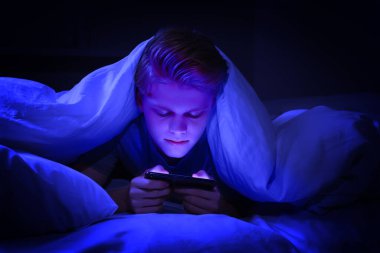 İnternet bağımlılığı. Genç çocuk gece yatakta battaniyenin altında akıllı telefon kullanıyor. Maviyle kaplanmış.