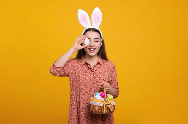 stock image Happy woman in bunny ears headband holding wicker basket of painted Easter eggs on orange background. Space for text