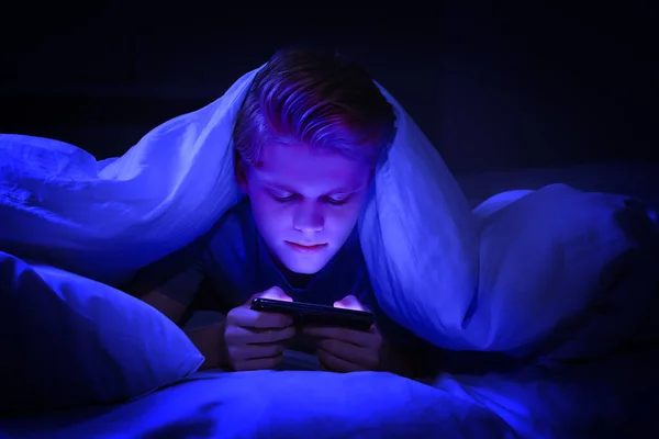 Internet addiction. Teenage boy using smartphone under blanket on bed at night. Toned in blue