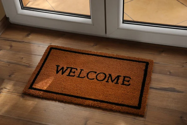 Door mat with word Welcome on wooden floor near entrance