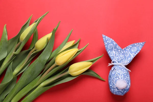 Easter bunny made of wrapping paper and egg near beautiful tulips on red background, flat lay