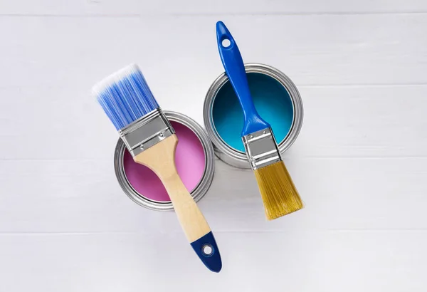 Cans of pink and light blue paints with brushes on white wooden table, flat lay