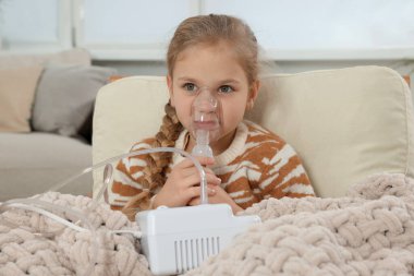 Evdeki koltukta nefes almak için nebulizör kullanan küçük kız.