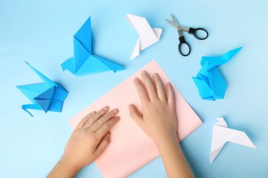 Origami art. Child folding paper on light blue background, closeup and top view
