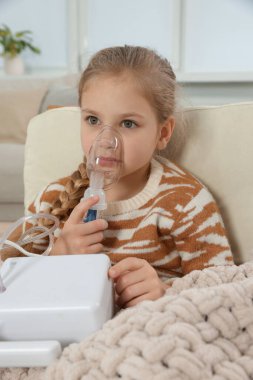 Evdeki koltukta nefes almak için nebulizör kullanan küçük kız.