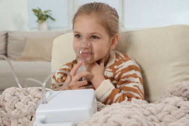 Evdeki koltukta nefes almak için nebulizör kullanan küçük kız.