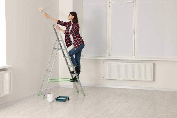 Young woman painting wall with roller on stepladder indoors. Room renovation