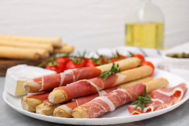 Plate of delicious grissini sticks with prosciutto, cheese and tomatoes on light grey table, closeup