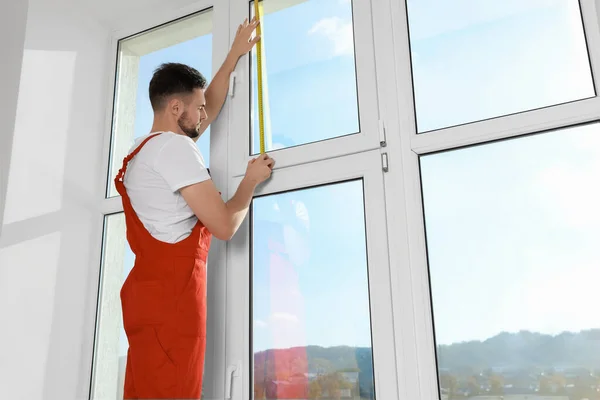 Worker in uniform measuring window with tape indoors. Roller blinds installation