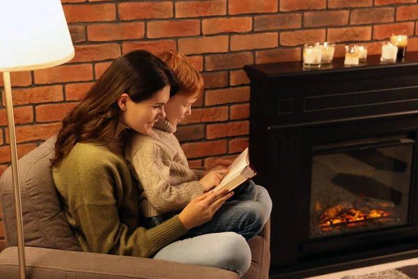 Happy mother and son reading book together near fireplace at home