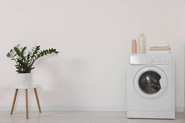 stock image Modern washing machine and potted plant on stool against white wall indoors