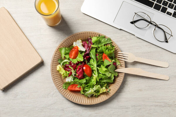 Fresh vegetable salad, glass of juice and laptop on white wooden table at workplace, flat lay. Business lunch