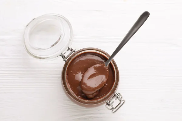 Jar with tasty chocolate paste and spoon on white wooden table, top view