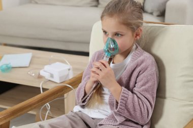 Evdeki koltukta nefes almak için nebulizör kullanan küçük kız.