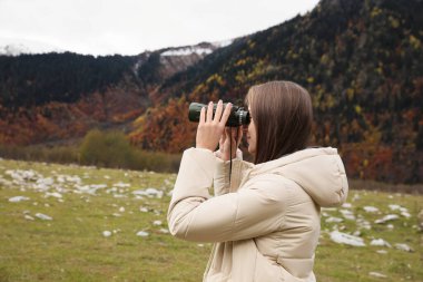 Woman looking through binoculars in beautiful mountains. Space for text