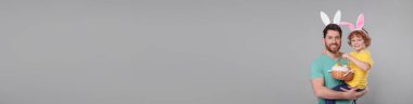 Mutlu baba ve oğlu. Tavşan kulaklı, açık gri arka planda paskalya yumurtası dolu sepet tutuyorlar. Mesaj için yer var. Pankart tasarımı