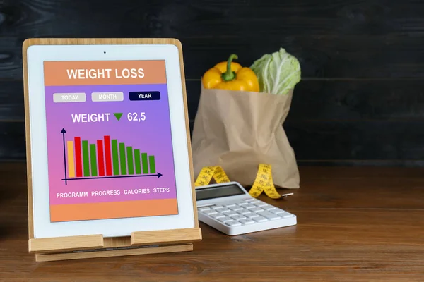 Tablet with weight loss calculator application and food products on wooden table, space for text