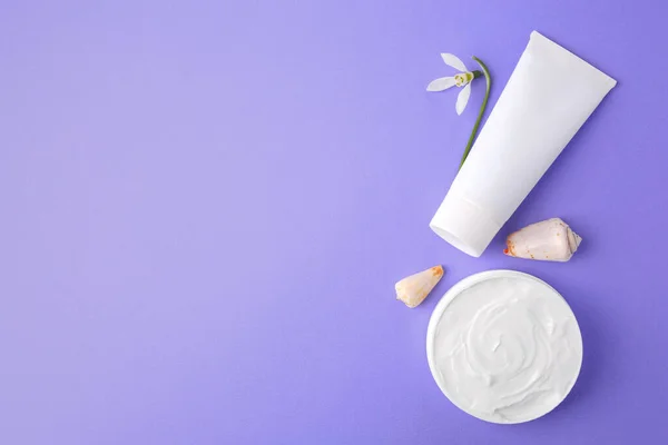 Jar and tube of cream, seashells with snowdrop flower on violet background, flat lay. Space for text
